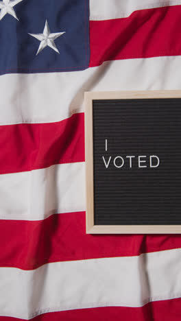 Vertical-Video-Of-Sign-Reading-I-Voted-Lying-On-US-Stars-And-Stripes-Flag-For-American-Election-2024